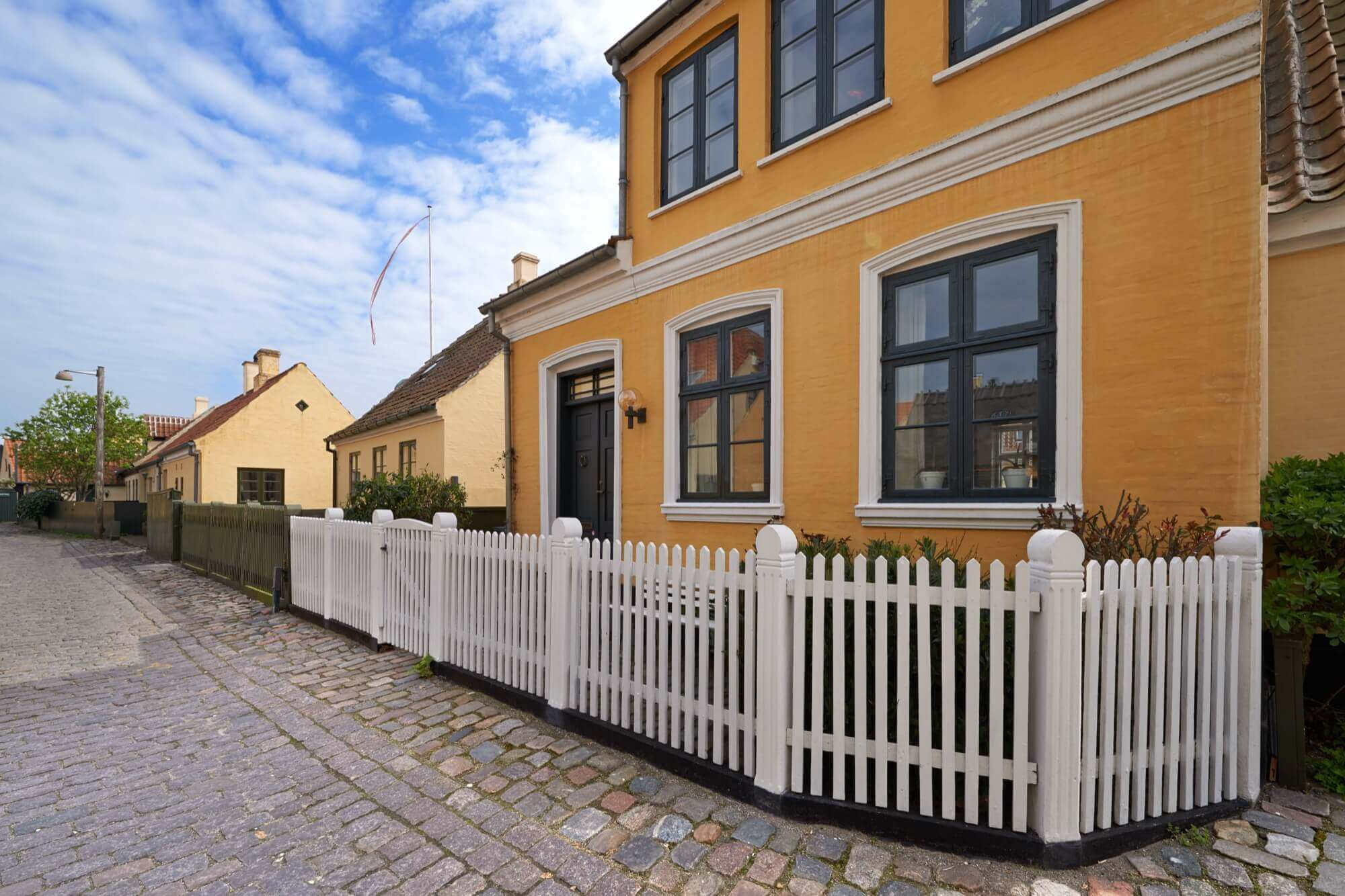 Gult hus med mörka fönsterkarmar och vitt staket på kullerstensgata, exempel på varm fasadmålning.