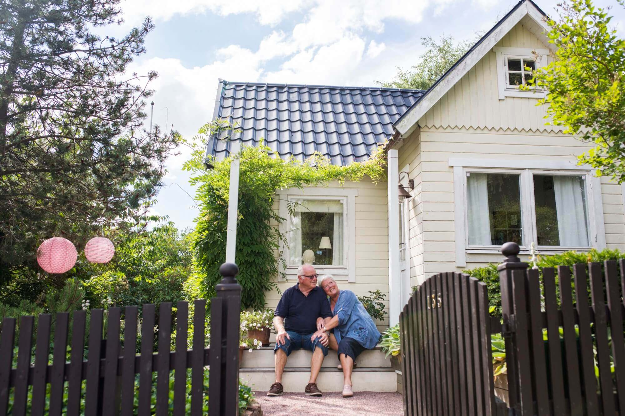 Äldre par sitter vid entrén till sin ljusa trästuga med blått tak och grönska runtom efter fasadmålning.