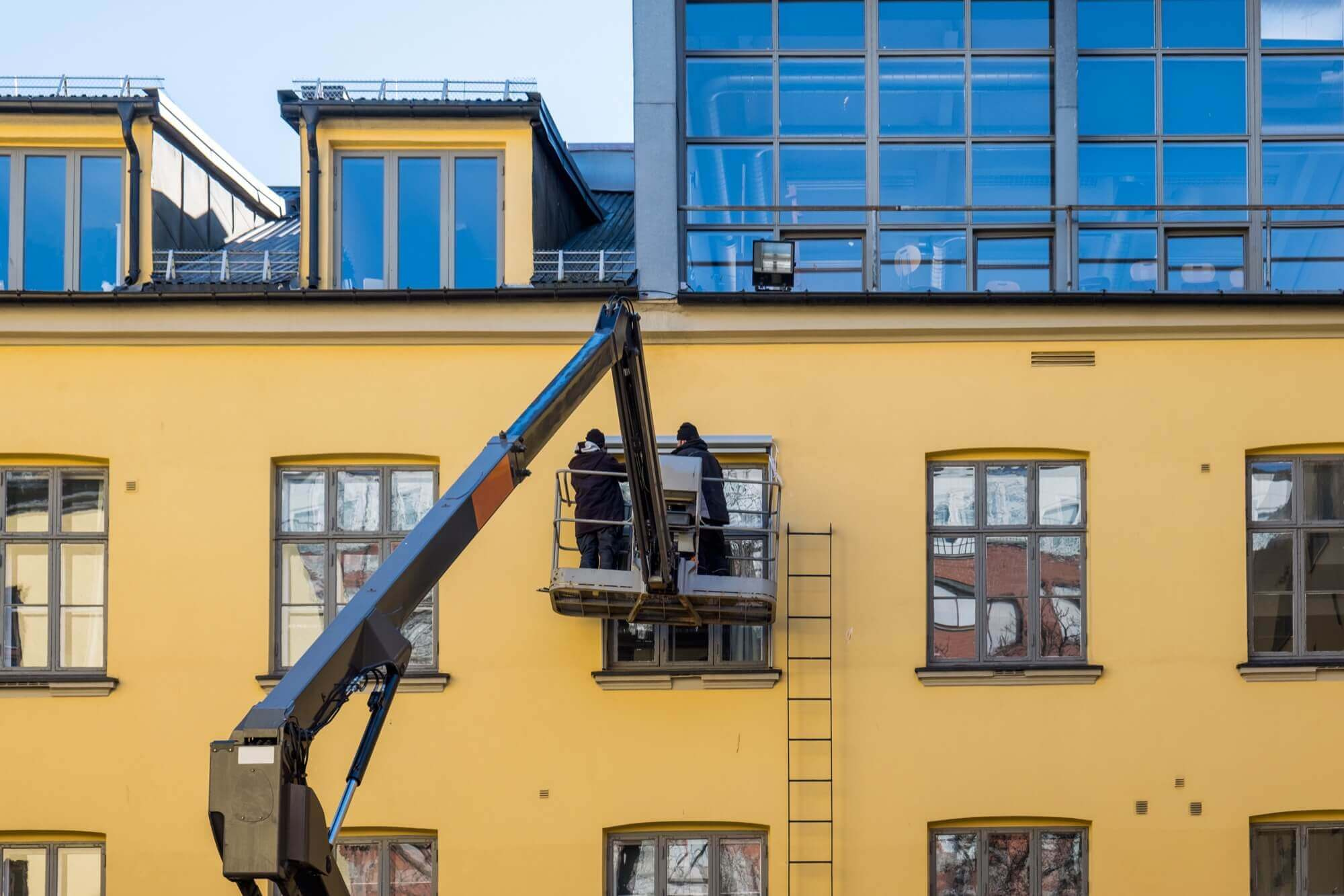 Arbetare på skylift underhåller fönster och utför fasadmålning på en gul byggnad.