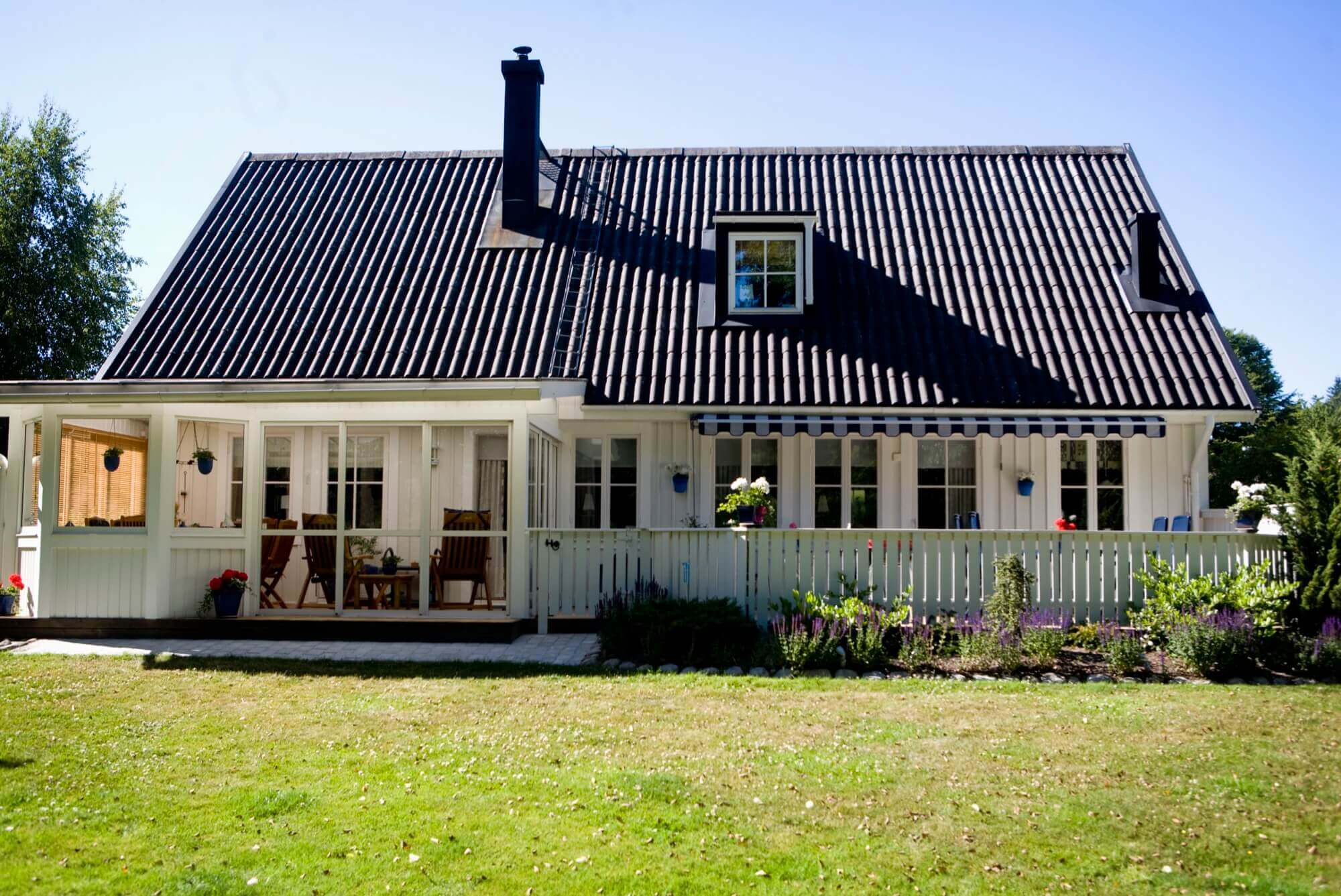 Vitmålad trävilla med stor veranda, svart tak och grönskande trädgård – välskött fasadmålning.